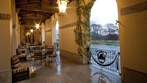 A covered outdoor seating area with tables and chairs, a view of a parking lot, and a railing with decorative ironwork. - Oheka Castle Hotel & Estate (Huntington)