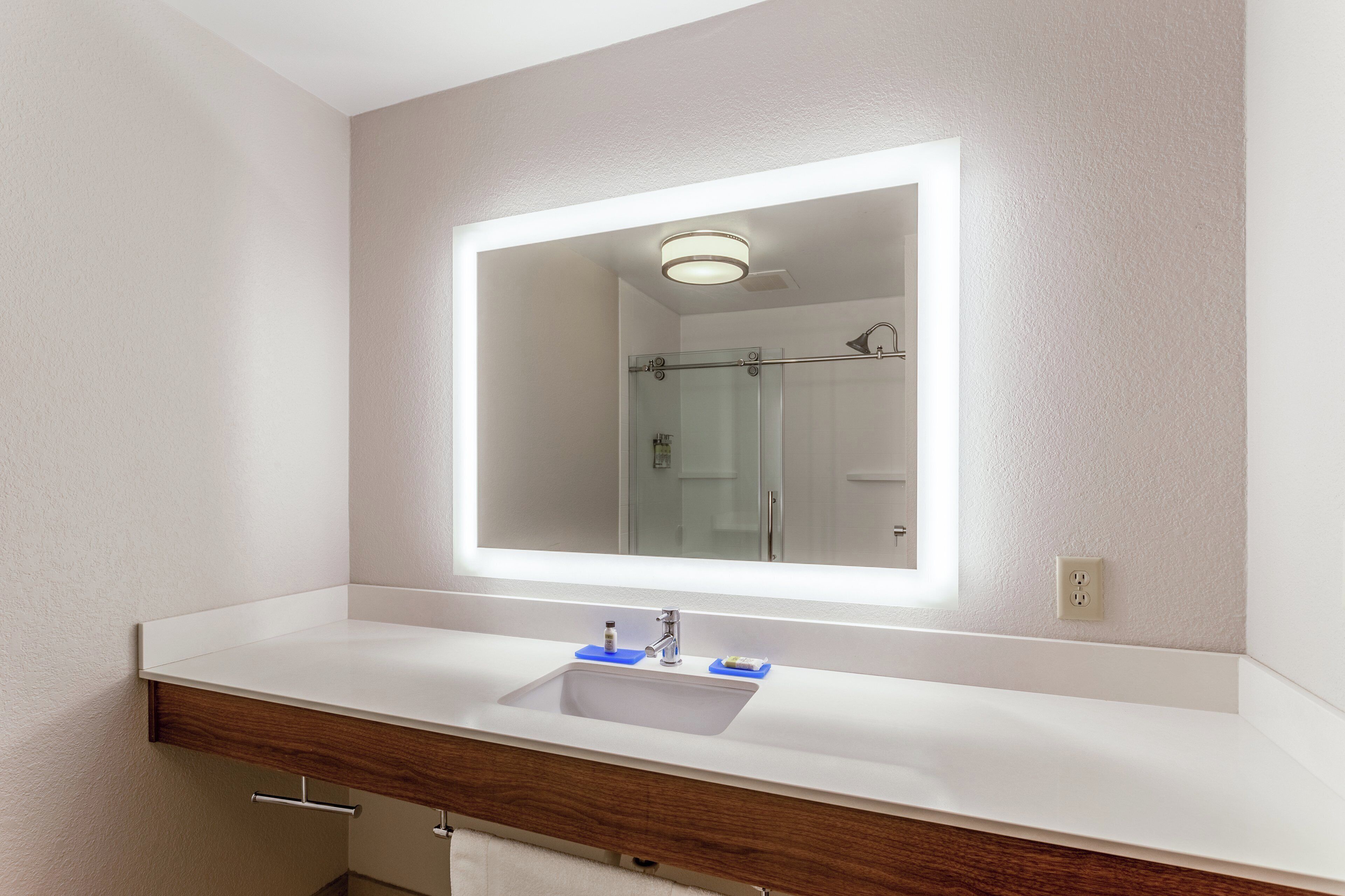 A bathroom with a white sink, a mirror with a light, and a shower visible in the reflection.