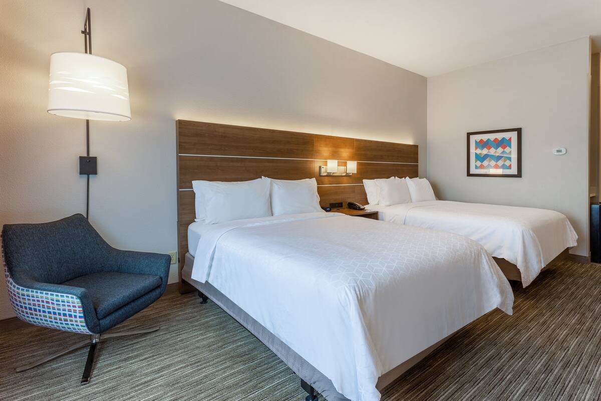 A hotel room with two beds, a chair, a lamp, and a wall-mounted light fixture.