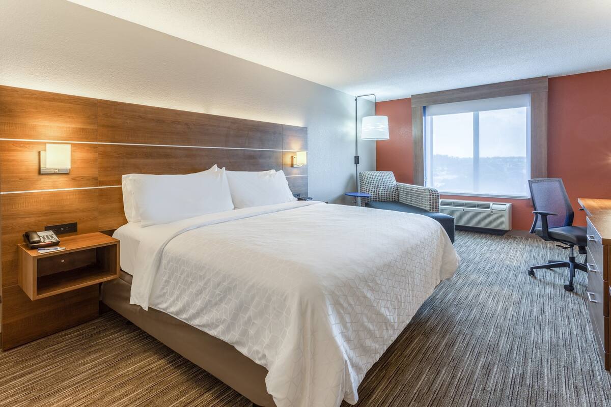 A hotel room with a large bed, a desk with a chair, a nightstand, and a window with a city view.