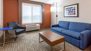 A modern hotel room with a blue sofa, a small table, a blue chair, a large window, and a framed artwork on the wall. - Holiday Inn Express & Suites O'Fallon/Shiloh by IHG (O'Fallon)