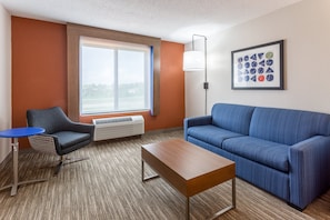 A modern hotel room with a blue sofa, a small table, a blue chair, a large window, and a framed artwork on the wall. - Holiday Inn Express & Suites O'Fallon/Shiloh by IHG (O'Fallon)