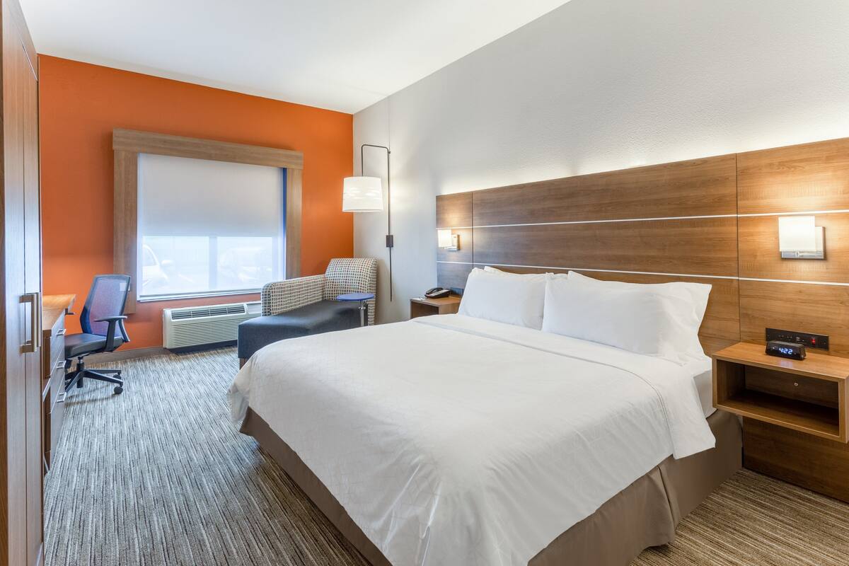 A hotel room with a large bed, a desk, a chair, a nightstand, and a wall-mounted lamp.