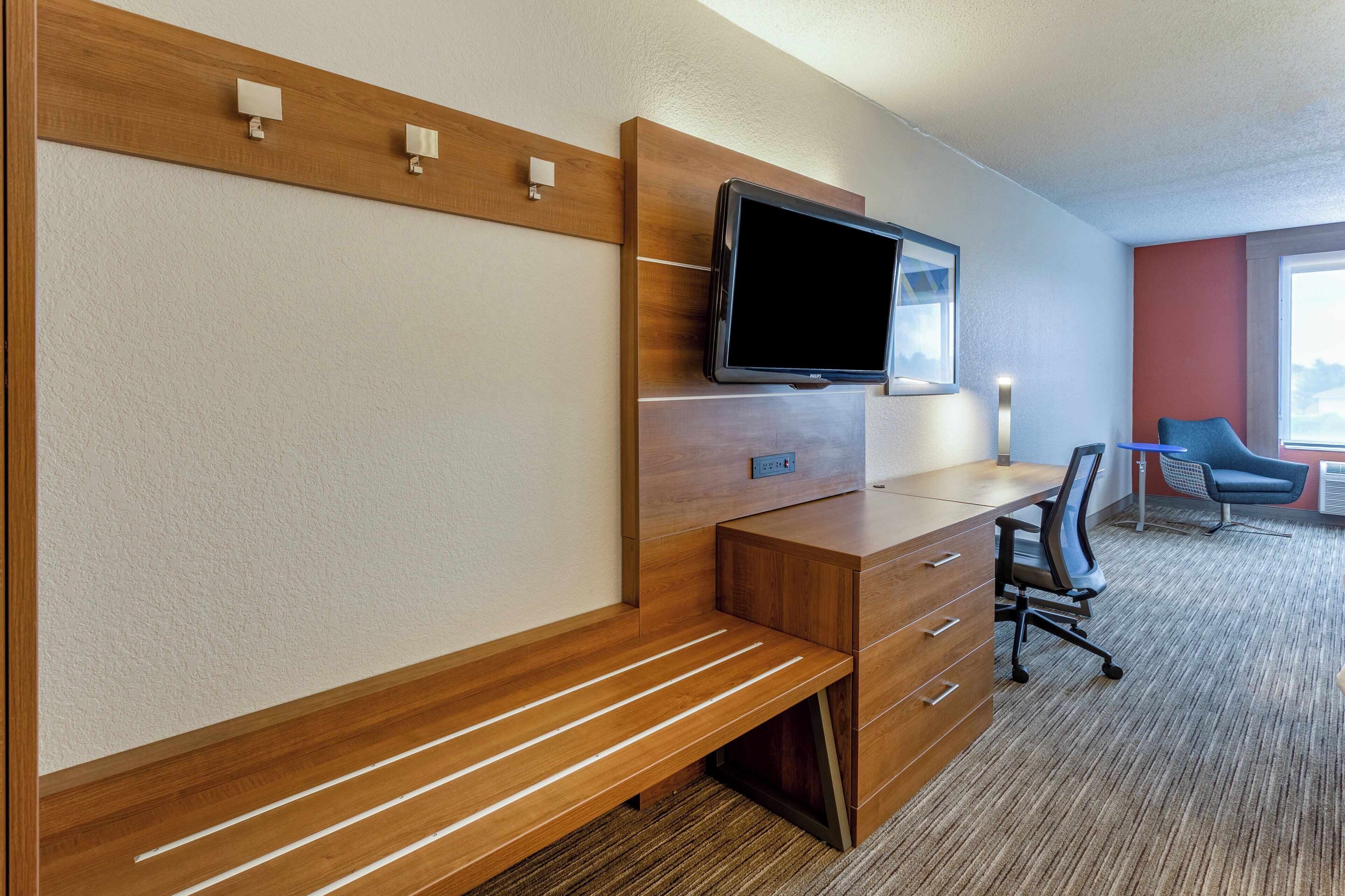 Habitación de hotel con un escritorio de madera, una televisión montada en la pared, una silla y un ventanal que proporciona luz natural.