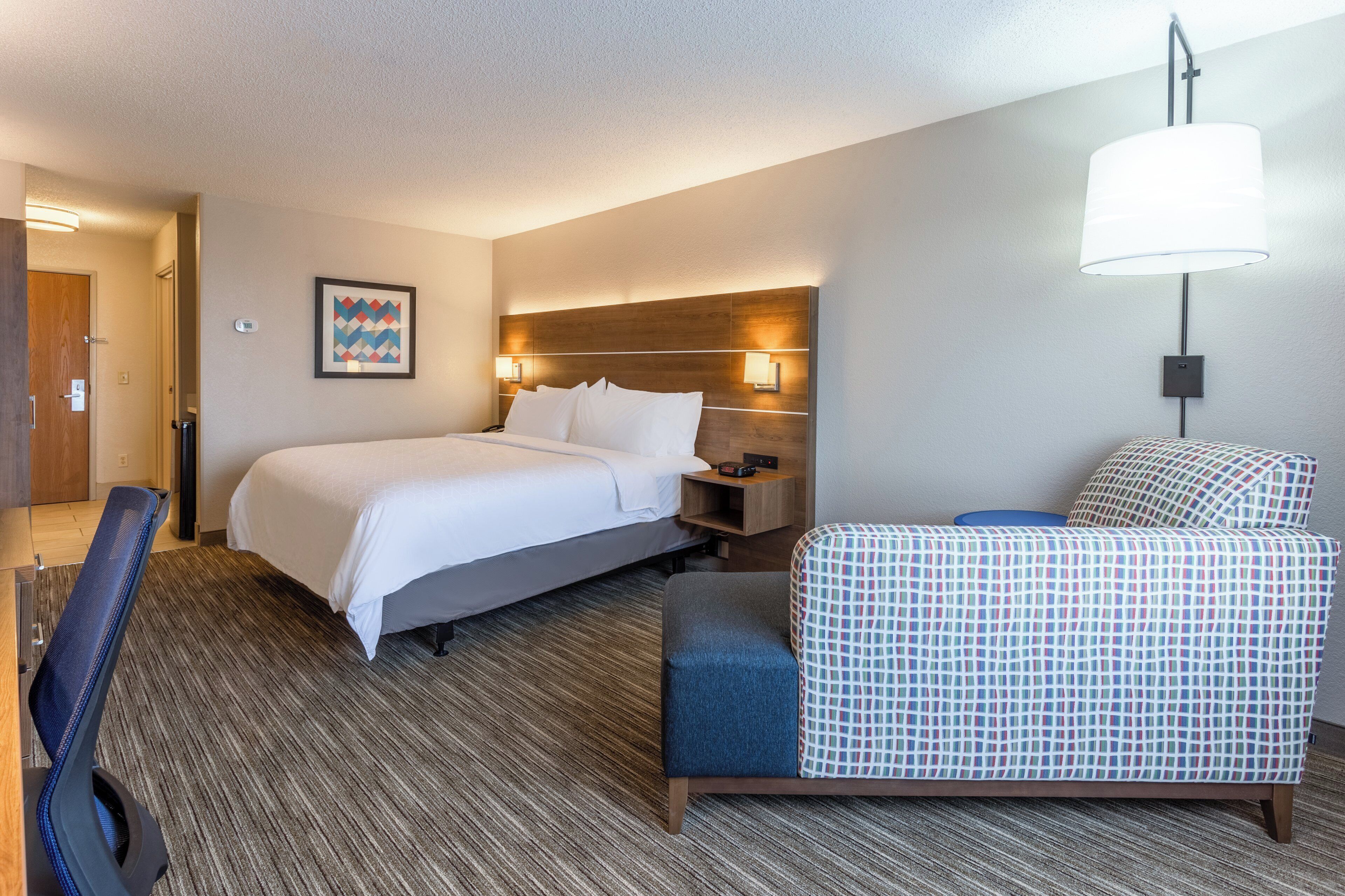 A hotel room with a bed, bedside table, lamp, chair, and a wall-mounted picture.