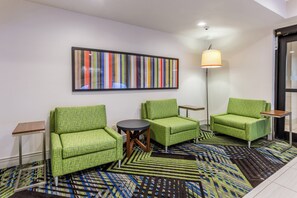A hotel room with green upholstered chairs, a small round table, a colorful abstract painting, a floor lamp, and a patterned carpet.