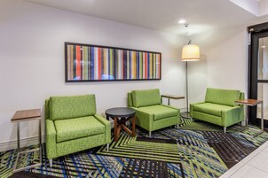 Habitación de hotel con sillas tapizadas en verde, una mesita redonda pequeña, un cuadro abstracto de colores, una lámpara de pie y una alfombra estampada.