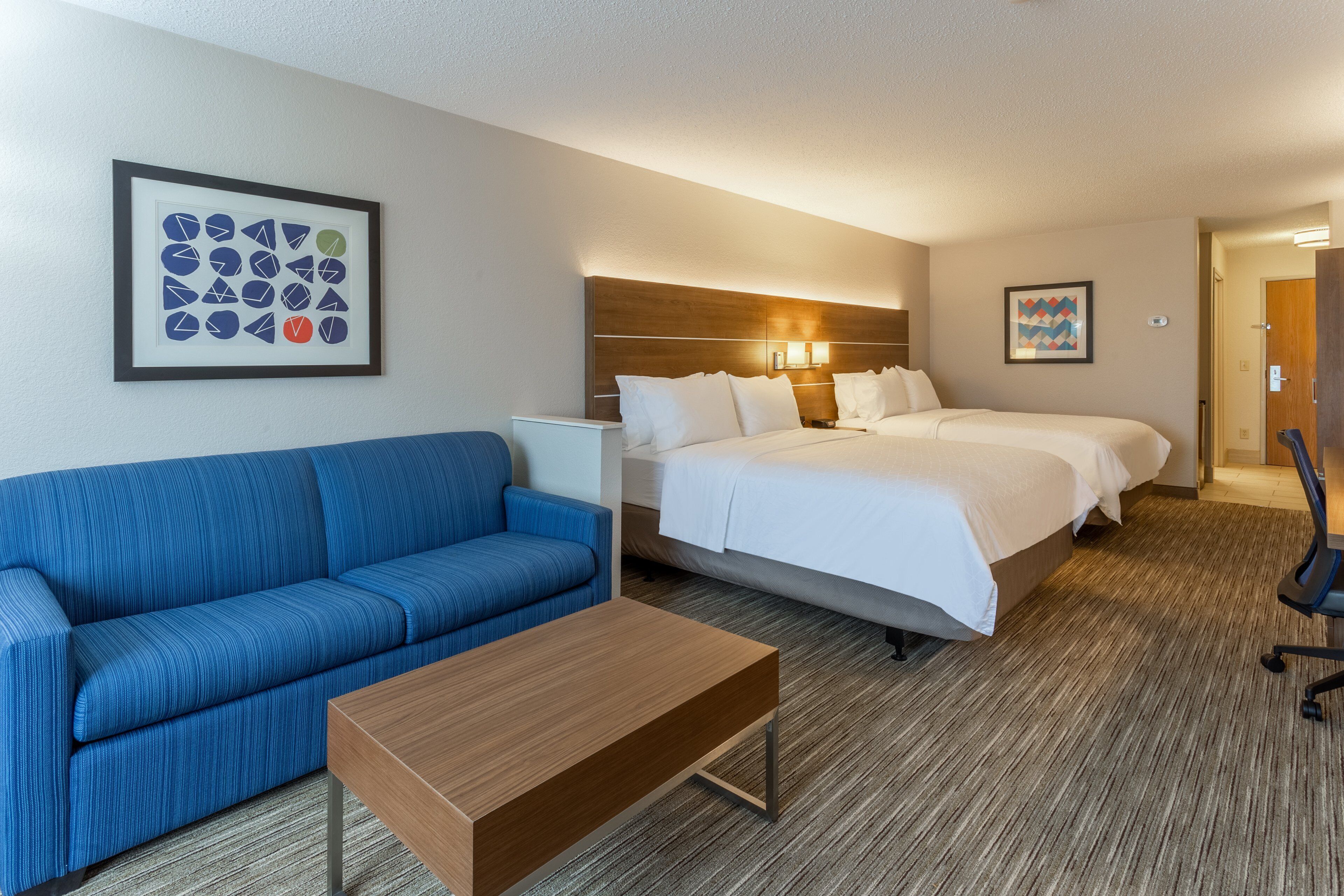 Habitación de hotel con sofá azul, una mesita de centro de madera, una cama con ropa de cama blanca y dos mesitas de noche.