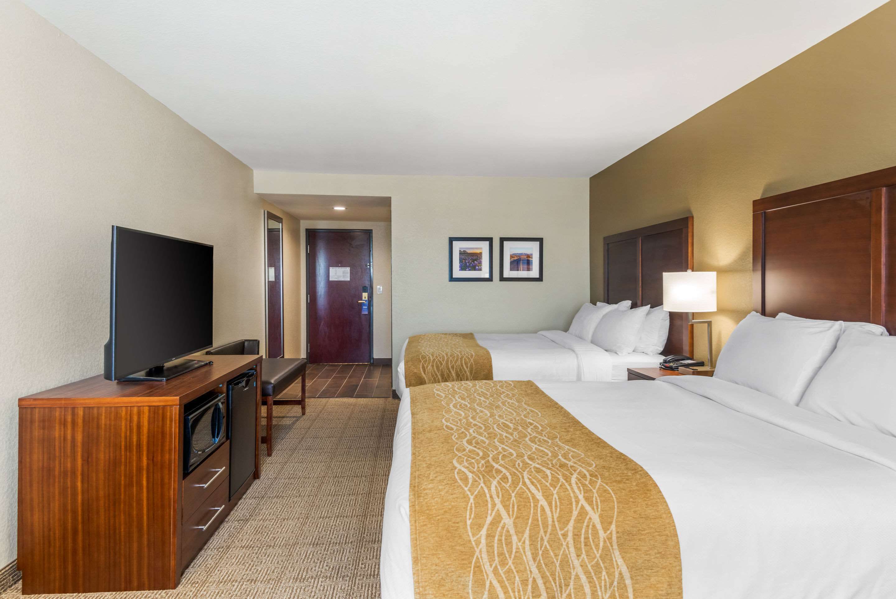 A hotel room with a TV, a desk with a computer, two beds, a bedside table with a lamp, and a door leading to a bathroom.