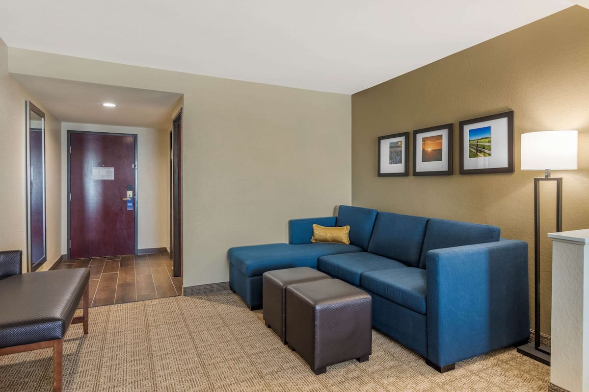 A modern hotel room with a blue sectional sofa, a small table, and a lamp. There are framed pictures on the wall and a door in the background.