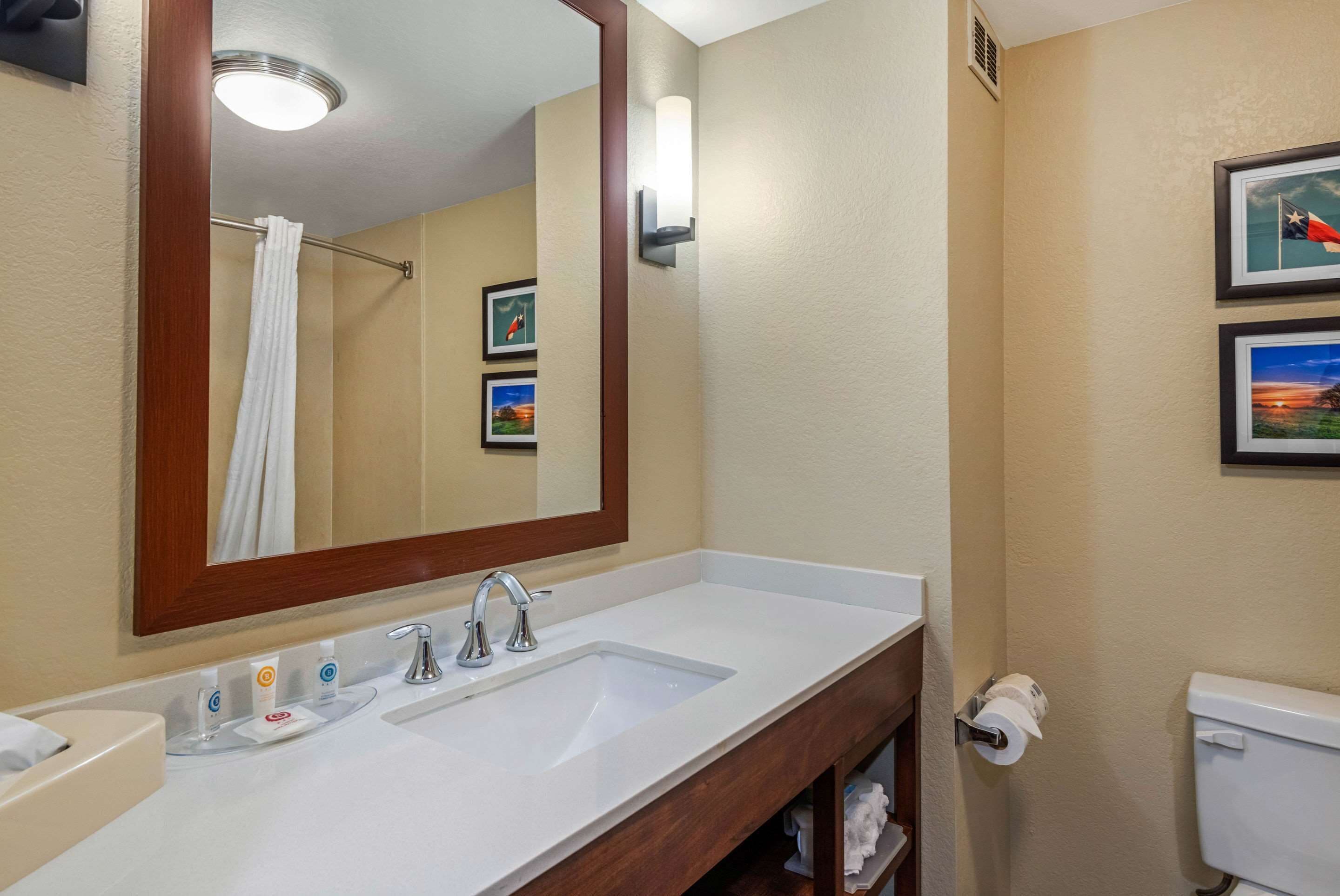 A hotel bathroom with a large mirror, a sink, a toilet, and framed pictures on the wall.