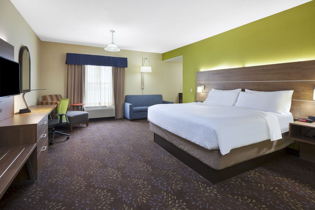 A hotel room with a large bed, a desk with a TV, a chair, a lamp, and a window with curtains.