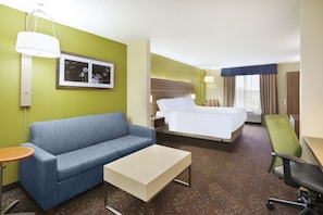 Habitación de hotel con una cama, un sofá azul, una mesa de centro de madera, una silla de oficina verde y una pequeña mesa redonda con una lámpara.