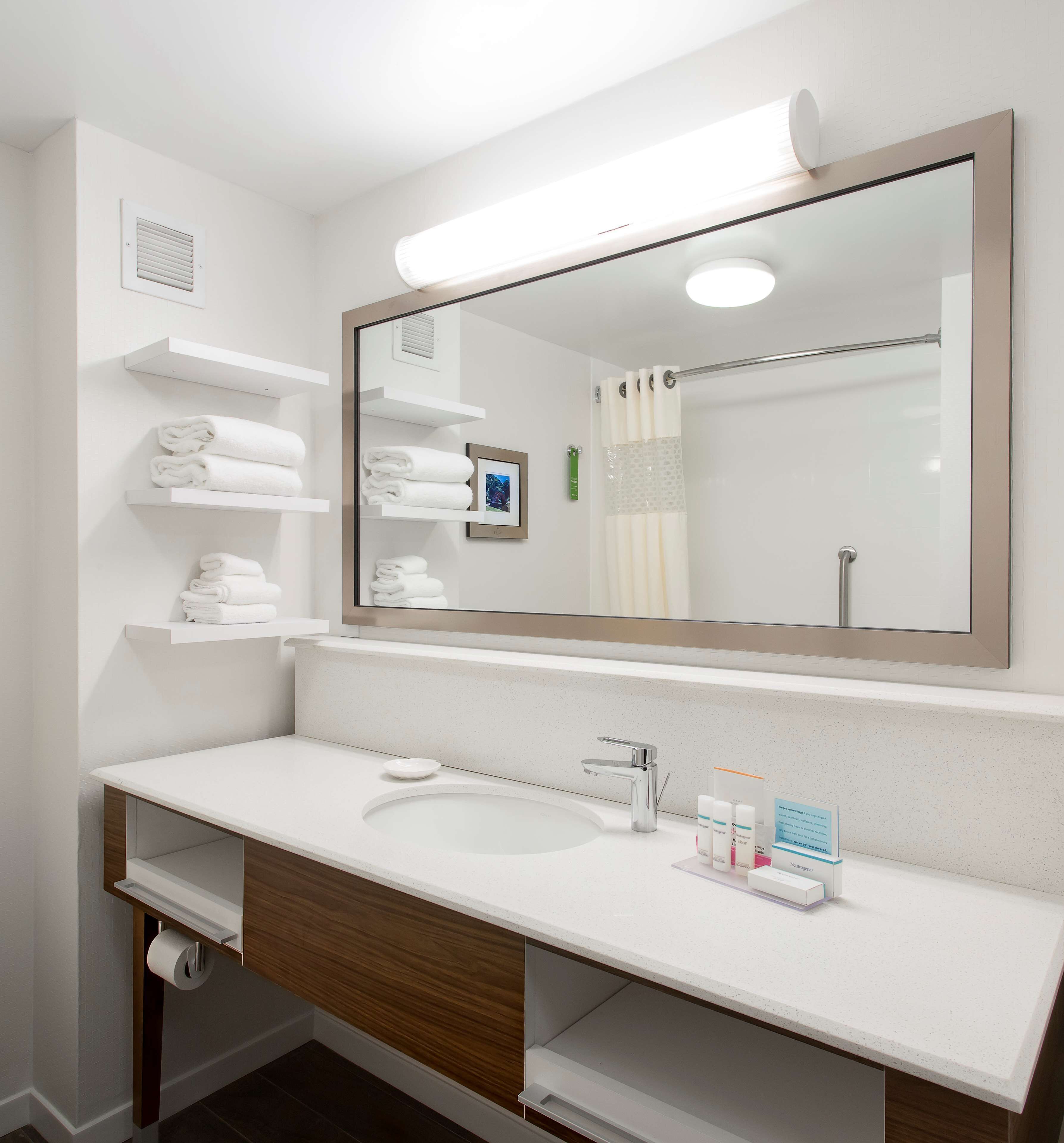A modern bathroom with a large mirror, white shelves with towels, a sink with a soap dispenser, and a toilet paper holder.