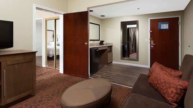 A hotel room with a kitchenette, a seating area with a sofa and ottoman, a television, and a bathroom visible through an open door. - Hampton Inn by Hilton Troy (Troy)