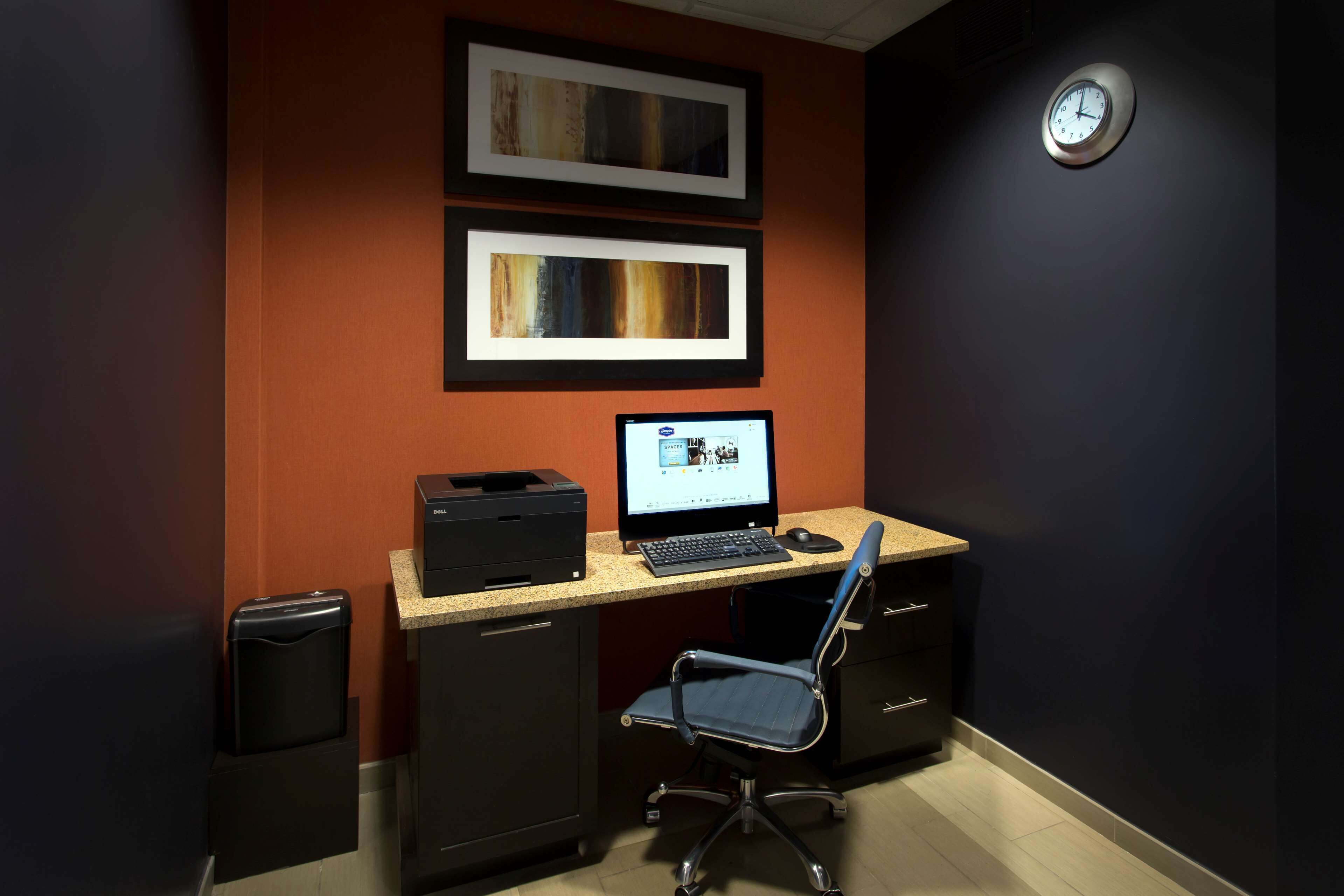 A home office setup with a desk, computer, chair, printer, and two framed pictures on the wall.