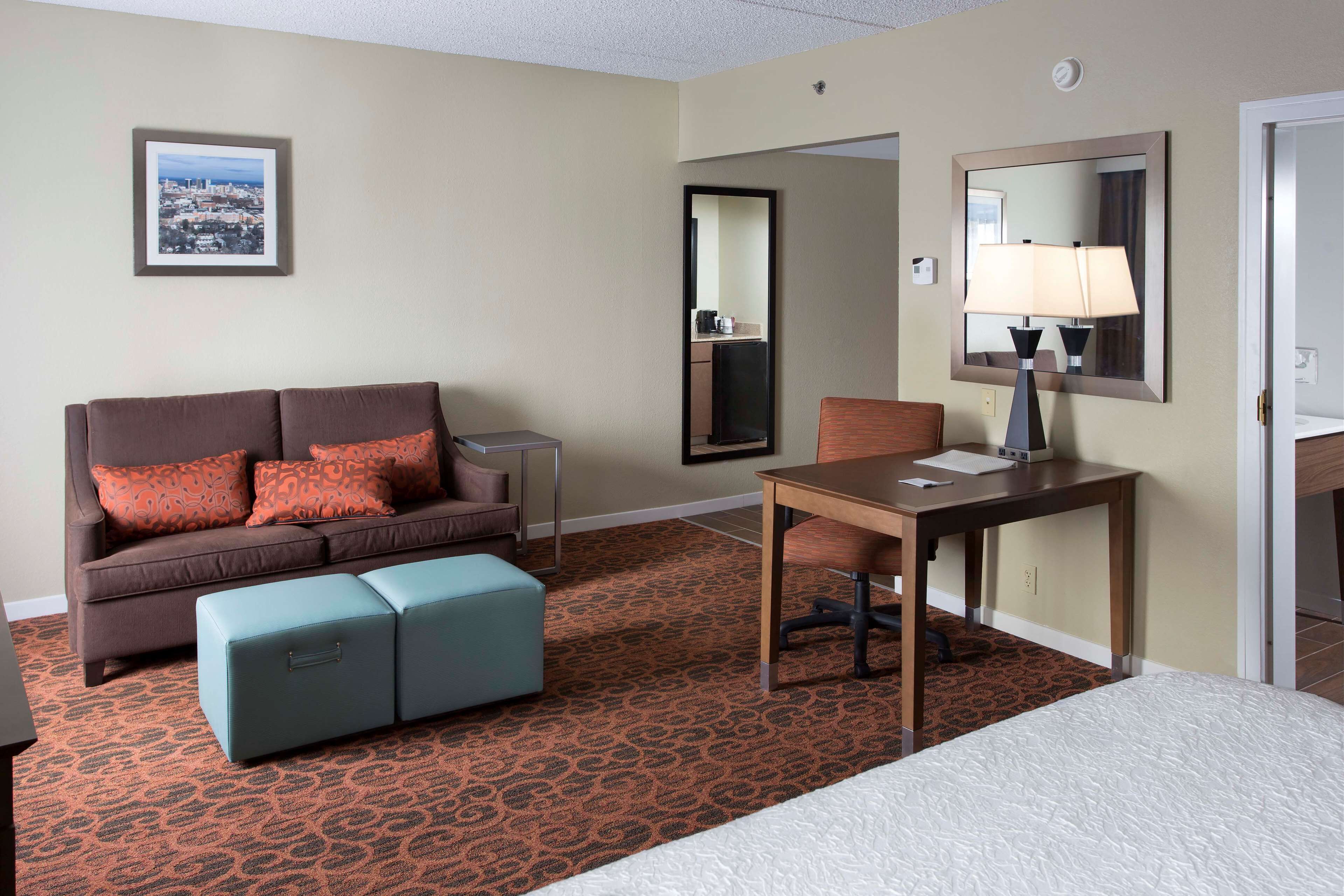 A hotel room with a brown sofa, a small table with a lamp, a blue ottoman, and a bed.