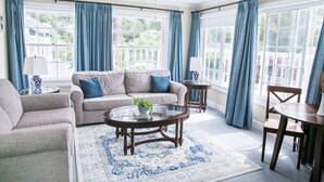 A spacious living room with a beige sofa, a round coffee table, and a wooden chair. There are large windows with blue curtains, a ceiling fan, and a view of greenery outside. - The Rigdon House (Cambria)