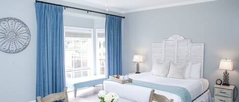 Un dormitorio con una cama, dos mesitas de noche, una mesa redonda de madera con sillas, un ventilador de techo y una ventana con cortinas.