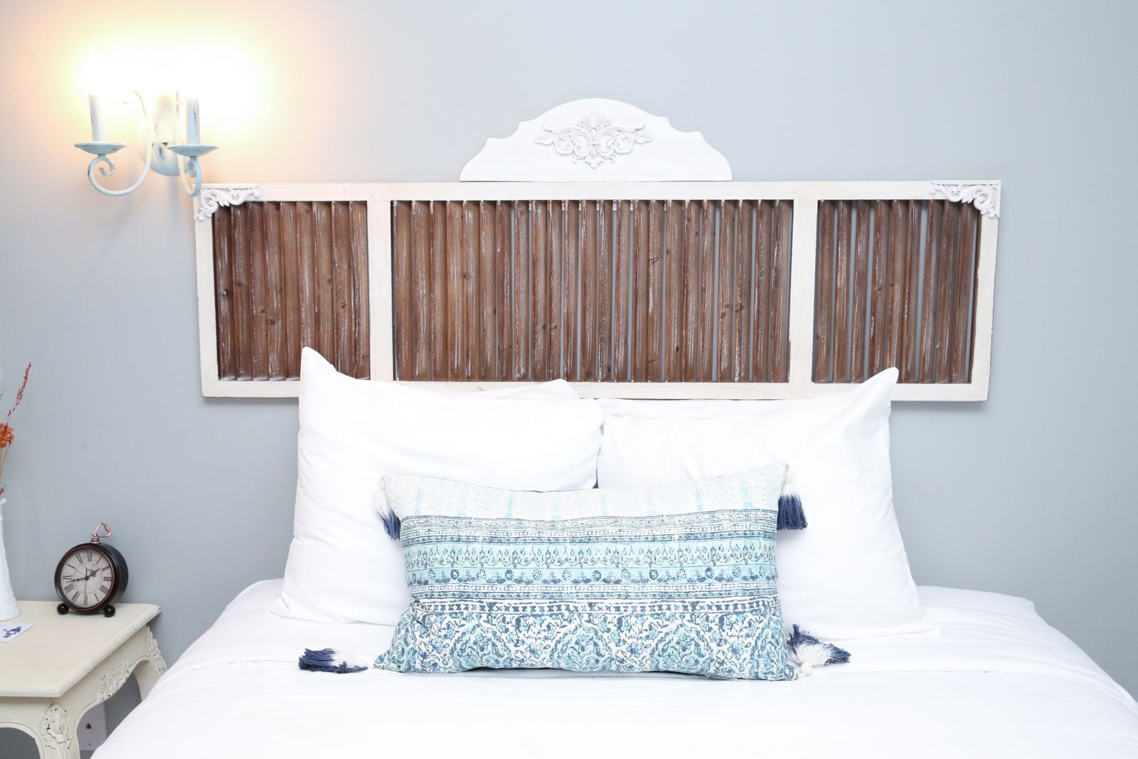 A bed with white bedding and patterned pillows, a wooden headboard, a wall-mounted light fixture, and a small table with a clock.
