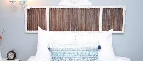 Een bed met wit beddegoed en gestreepte kussens, een houten hoofdbord, een wandlamp en een klein tafeltje met een klok.