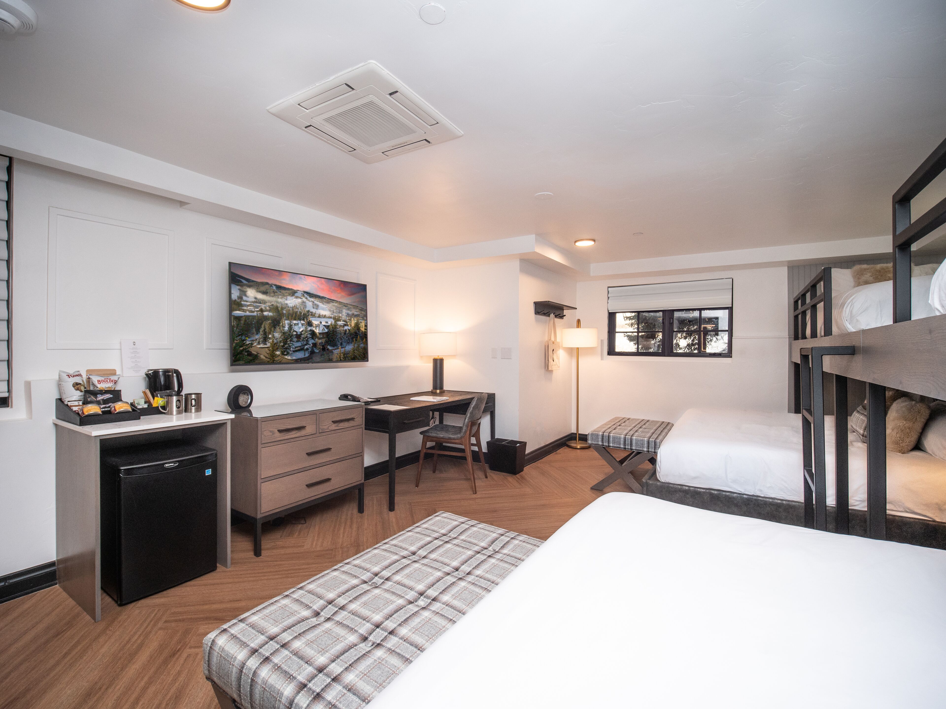 A hotel room with a bed, a desk, a chair, a small fridge, a lamp, and a window.