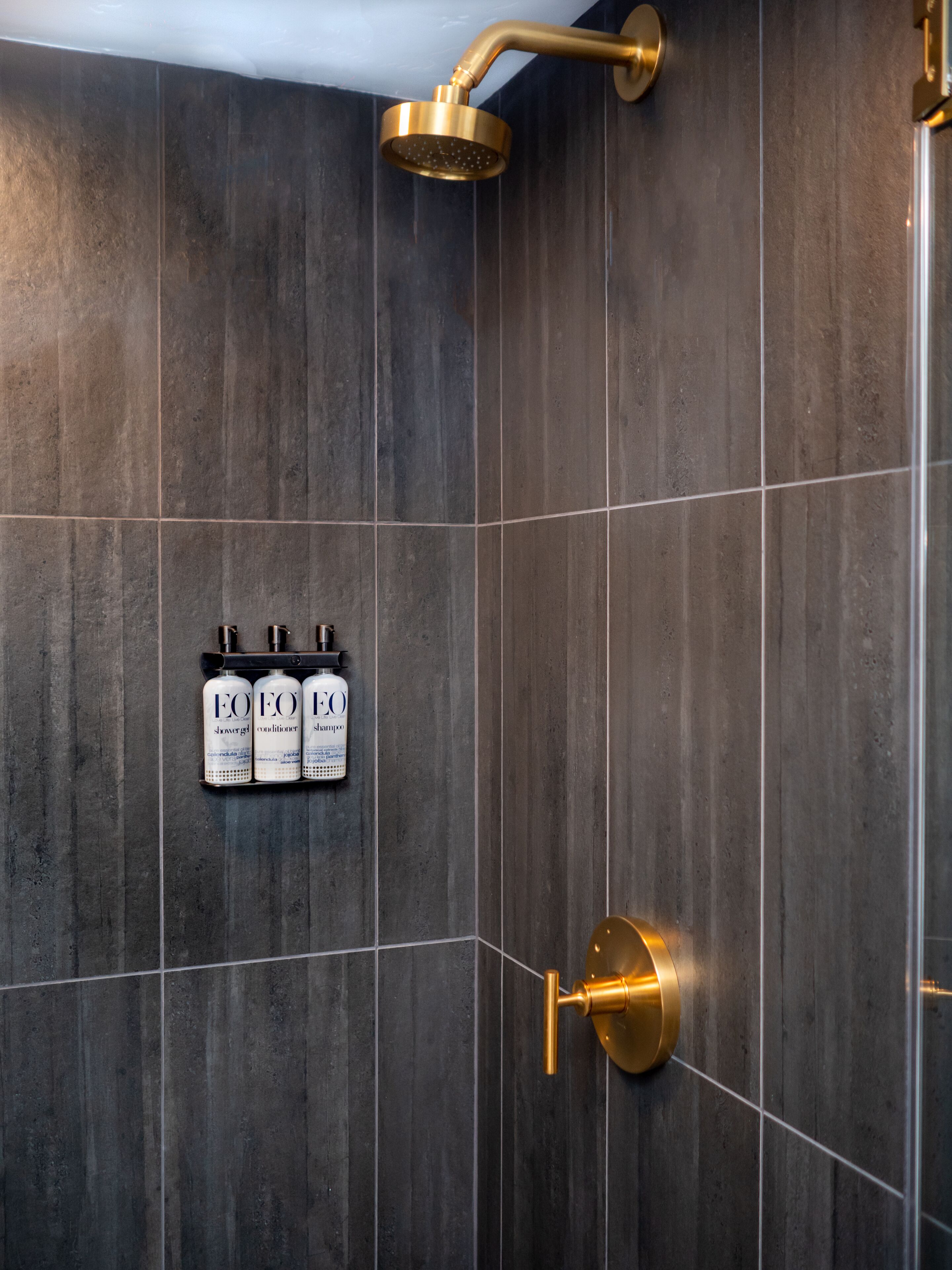 A modern bathroom with dark tiles, a gold showerhead, and three bottles of EFOX products.