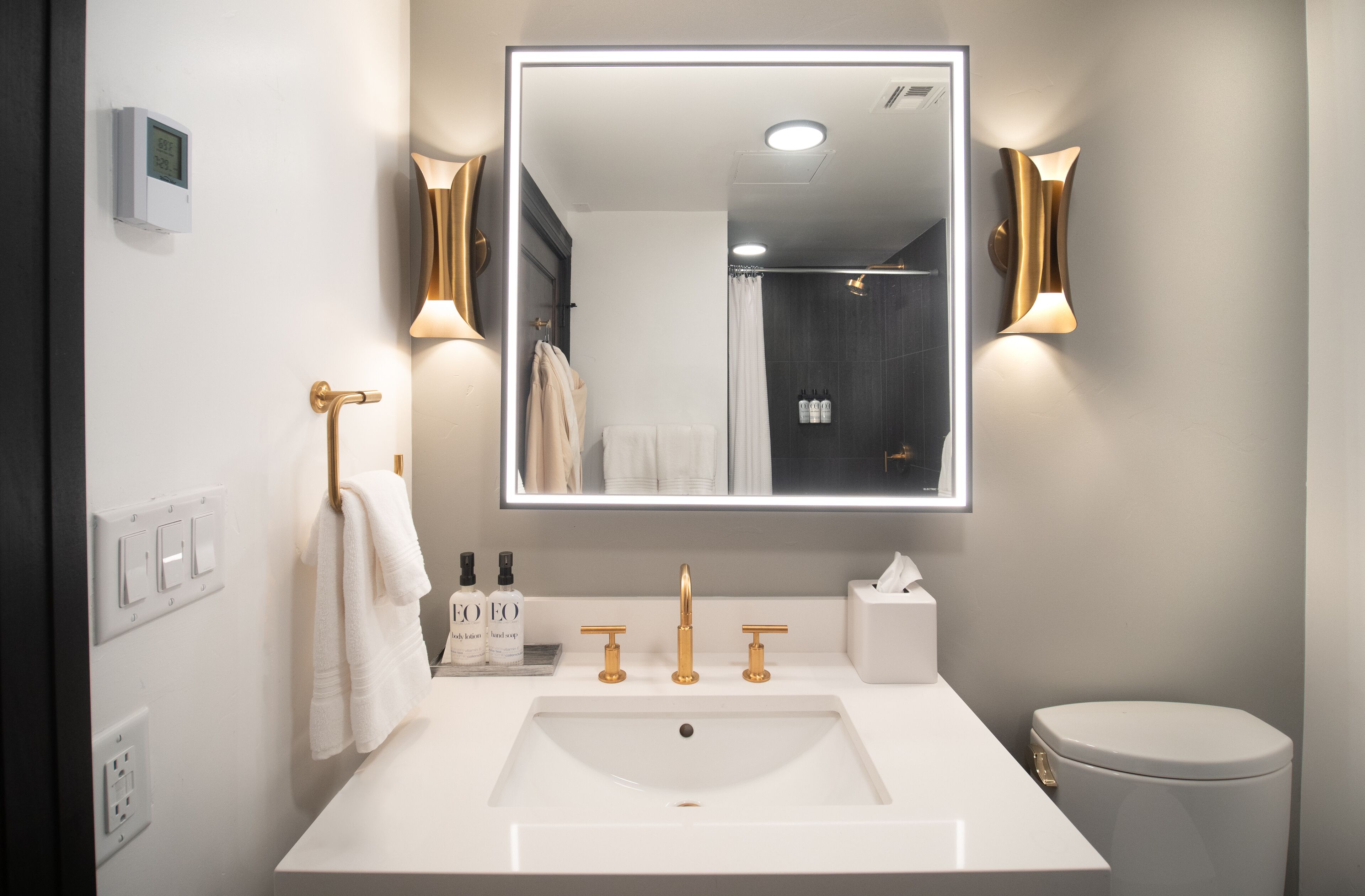 Ein modernes Badezimmer mit großem Spiegel, weißem Waschbecken, goldenen Armaturen und Toilette.