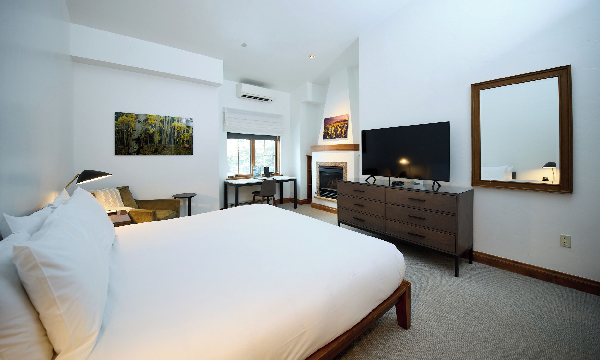 A hotel room with a large bed, a desk with a chair, a television, a fireplace, and a large mirror.