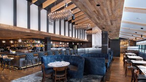 Un espace bar moderne avec un plafond haut, des poutres en bois et un comptoir avec des tabourets. On y trouve également des coins salon avec des chaises et des tables à revêtement bleu.