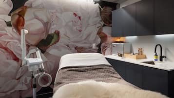 A bedroom with a large floral mural, a bed with a fluffy white cover, a bedside table with a lamp, and a small wooden box with toiletries.
