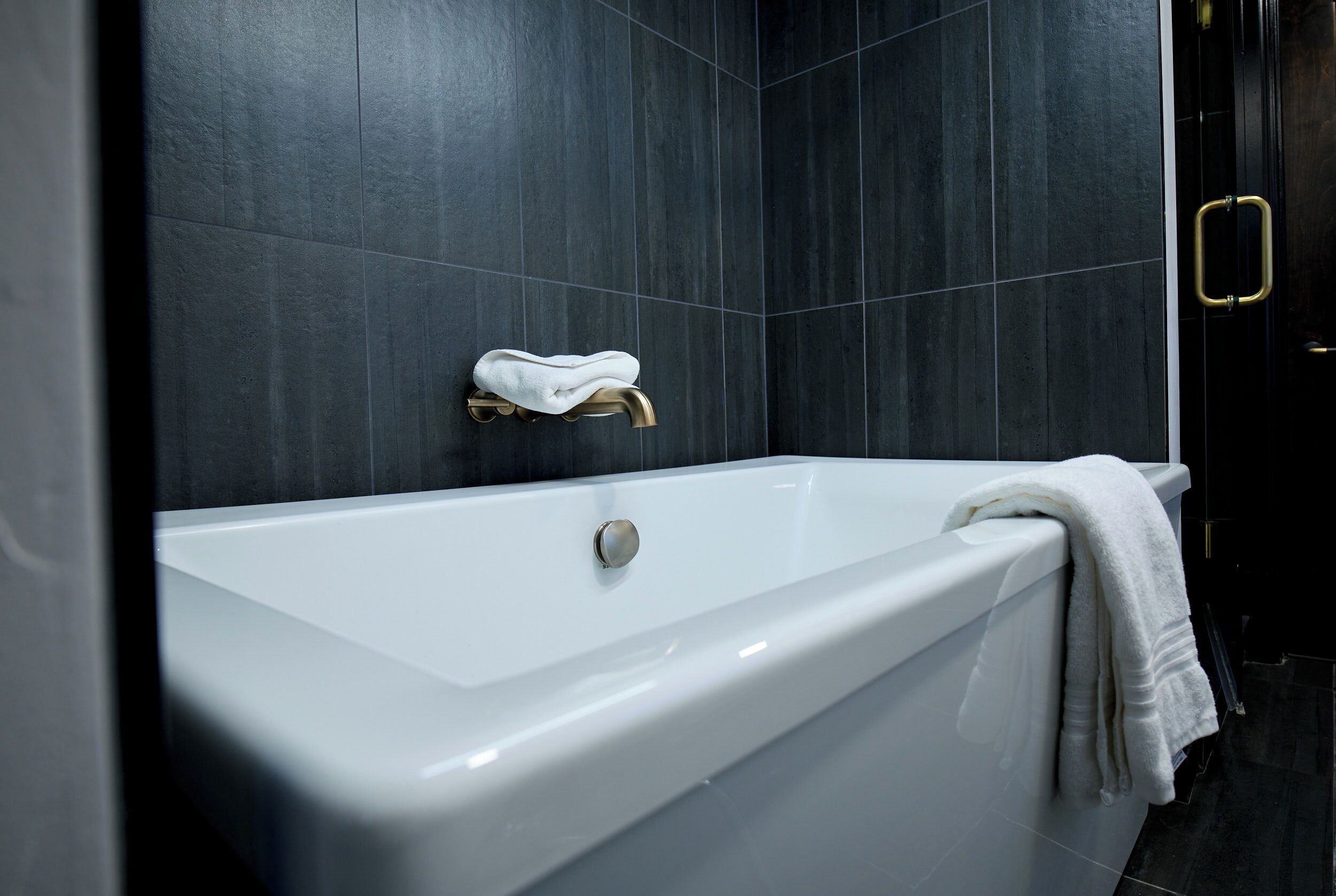 Ein modernes Badezimmer mit einer weißen Badewanne, dunkel gefliesten Wänden und einer goldenen Armatur.