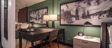 Habitación de hotel con escritorio, silla y una gran foto enmarcada de un paisaje montañoso nevado.