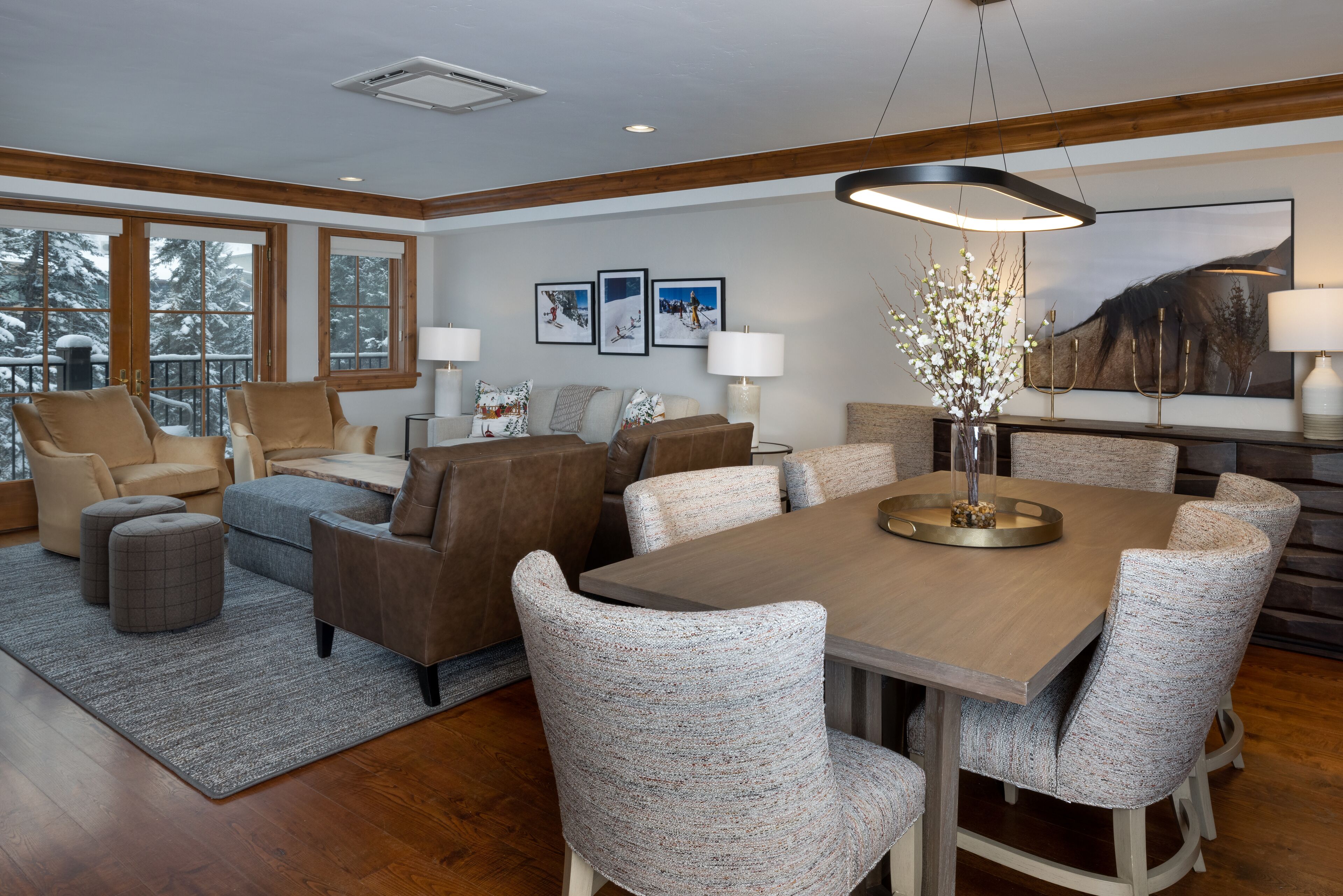 A modern living room with a wooden dining table, grey upholstered chairs, a grey rug, and a large window with a view of snow-covered trees.