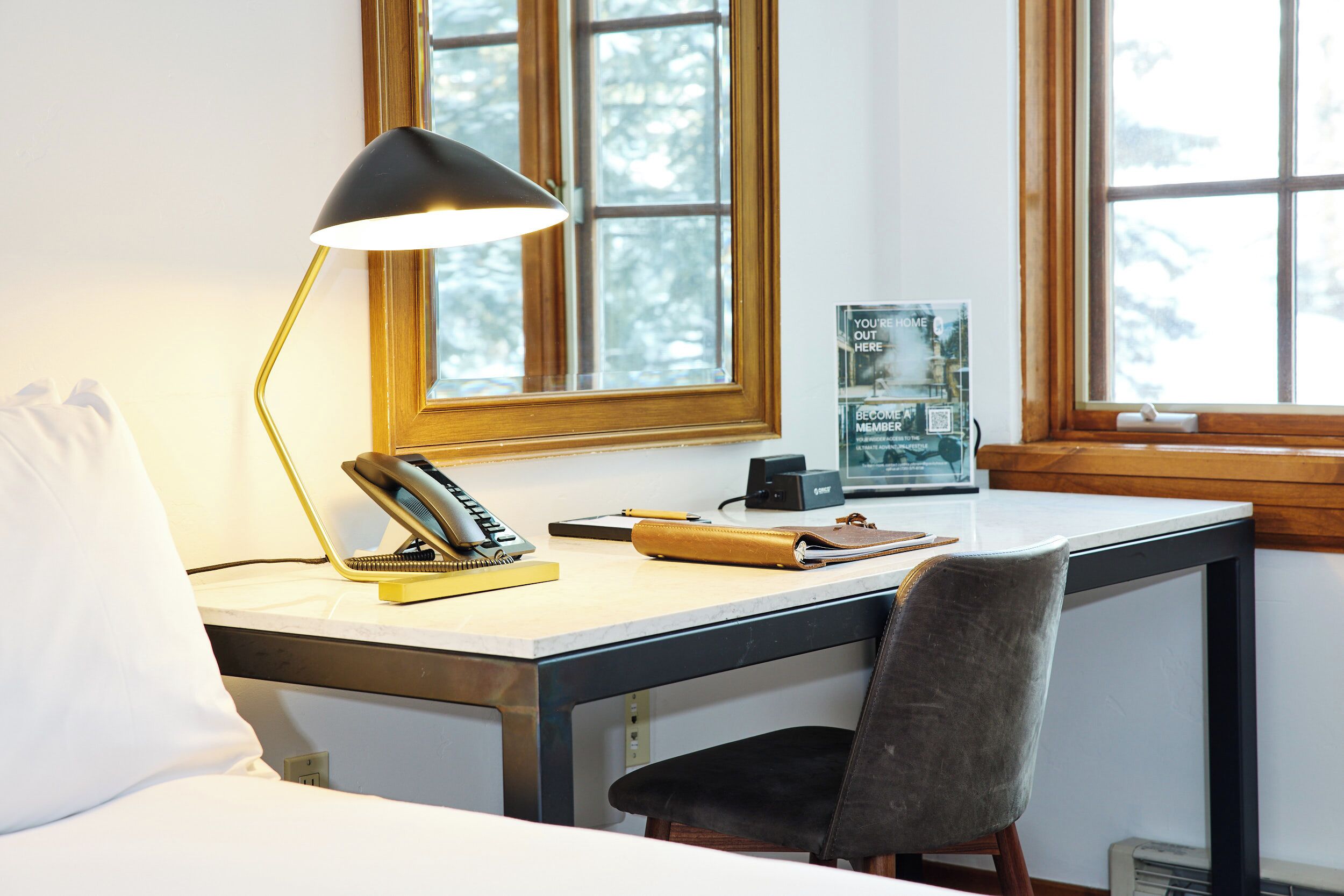 A room with a desk, a lamp, a chair, a bed, and a window with a view of trees.