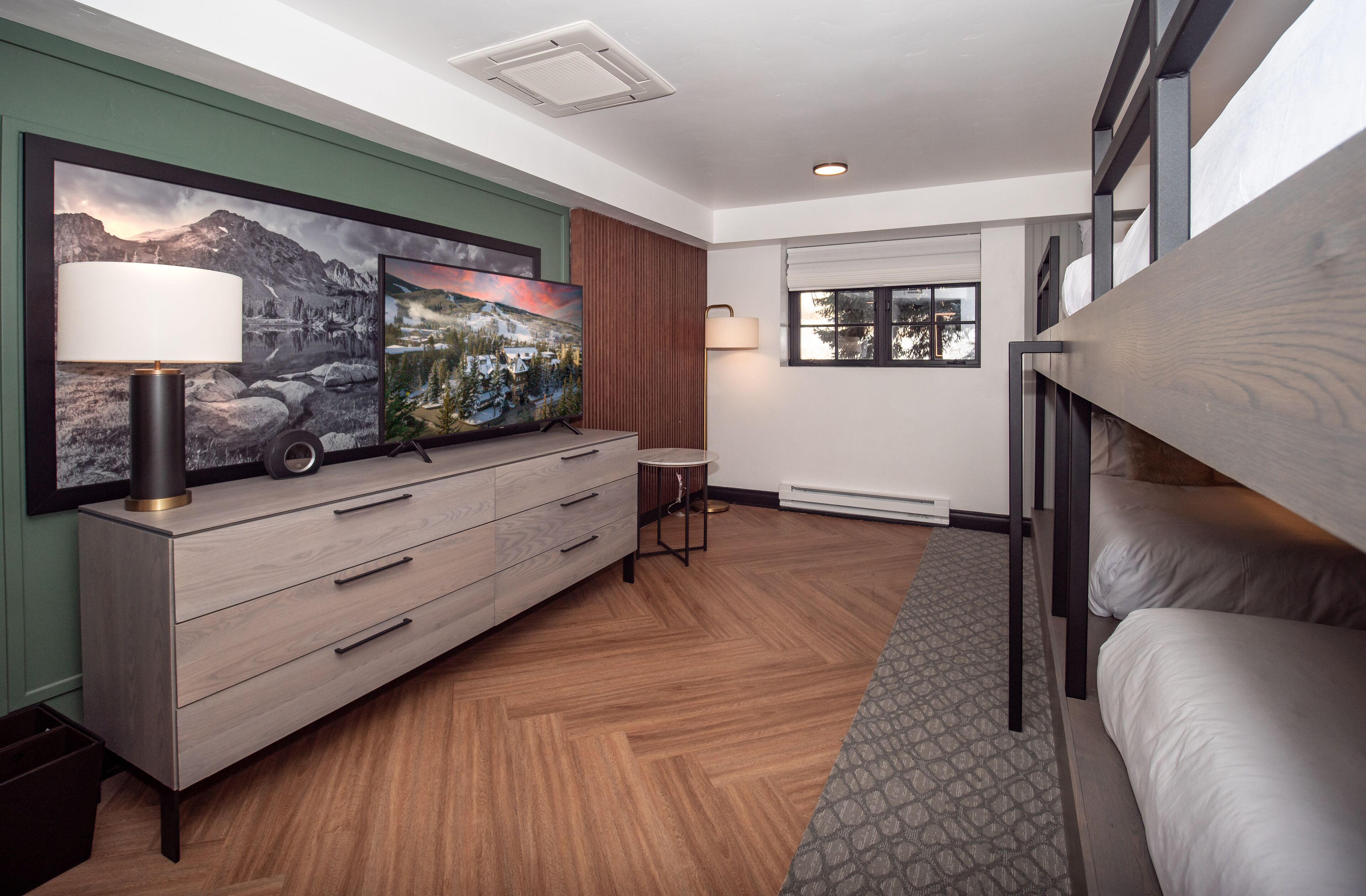A modern hotel room with a bunk bed, a wooden dresser with a lamp, a framed landscape painting, and a window with a view of trees.