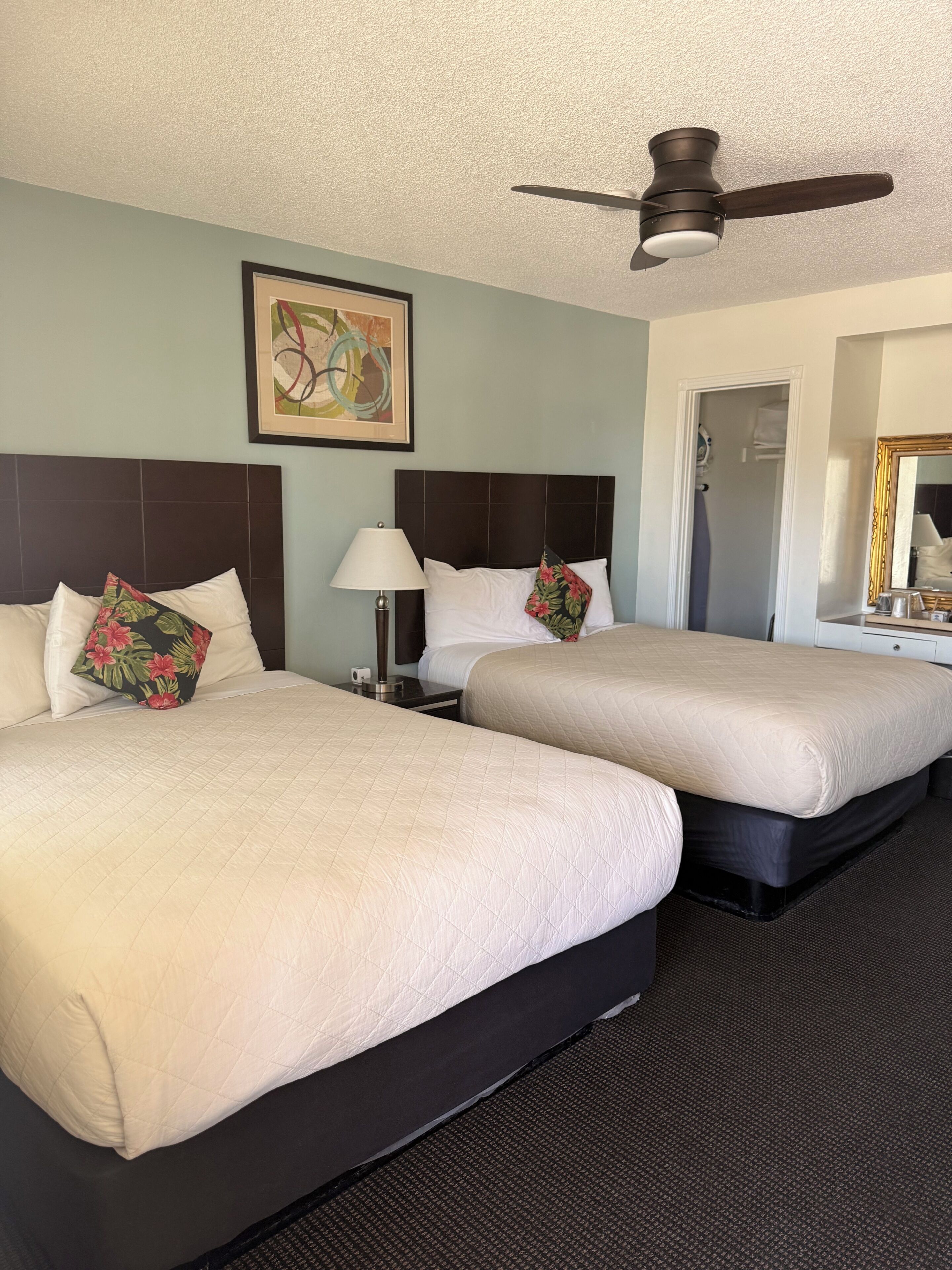 A hotel room with two beds, a ceiling fan, a framed artwork, a nightstand, and a mirror.