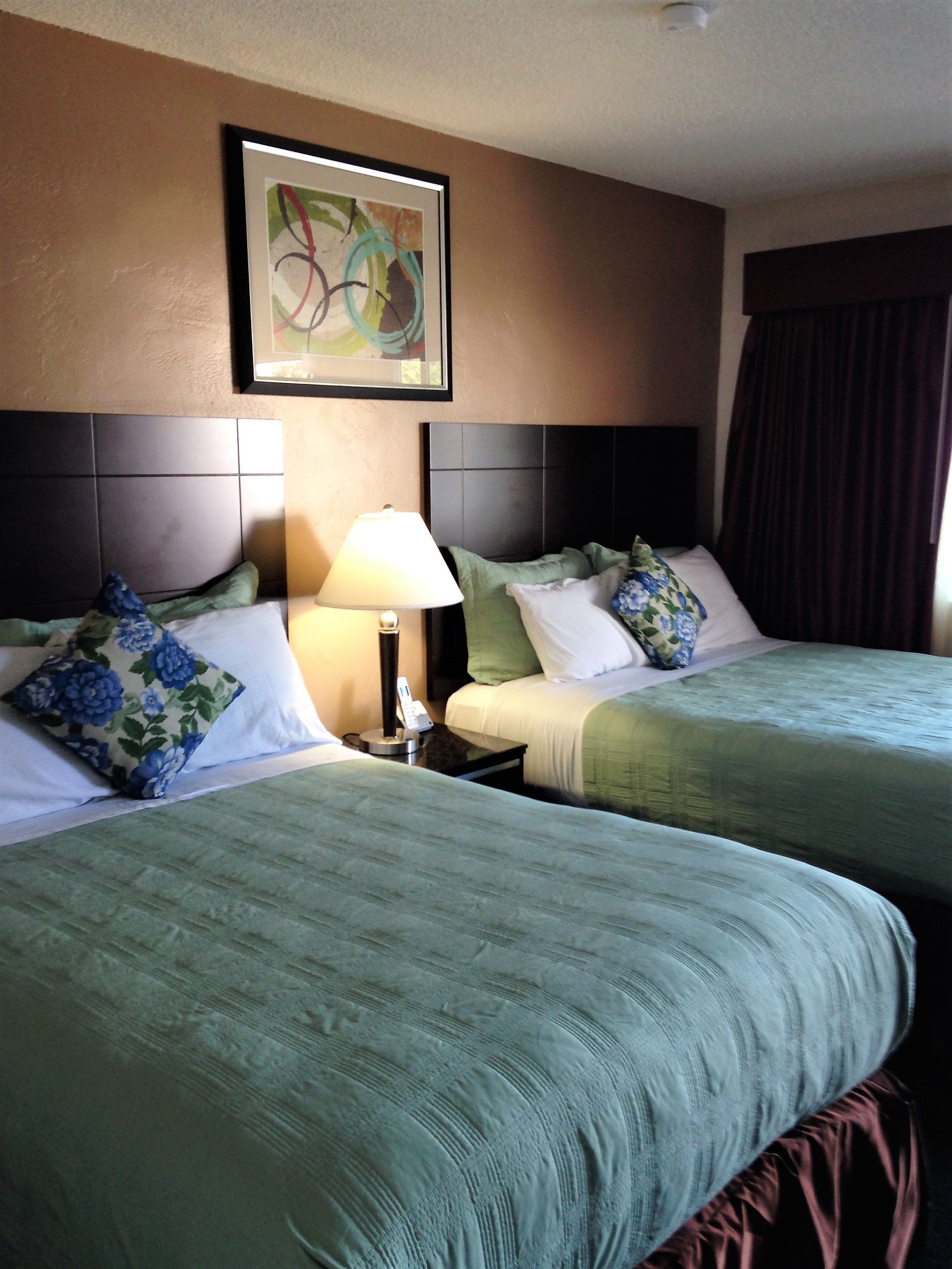 Quarto de hotel com duas camas, uma mesa de cabeceira com luminária, um quadro abstrato emoldurado na parede e uma janela com cortinas.