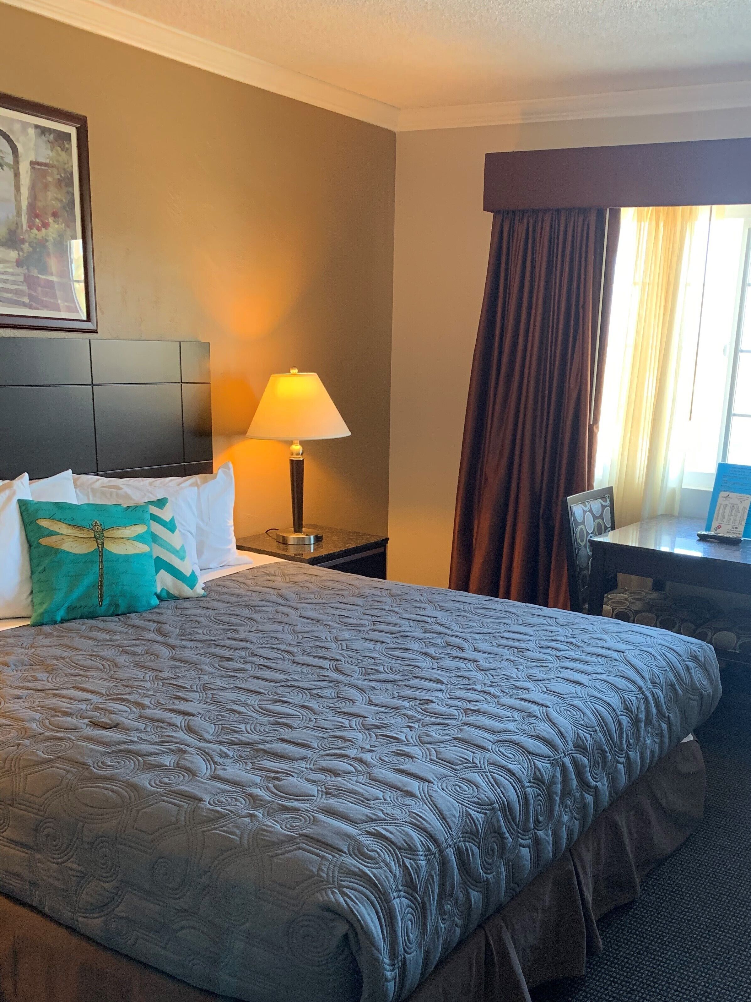 A hotel room with a large bed, a nightstand with a lamp, a desk with a chair, and a window with curtains.
