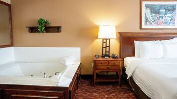 A hotel room with a large bathtub, a bedside table with a lamp, and a bed with white linens.