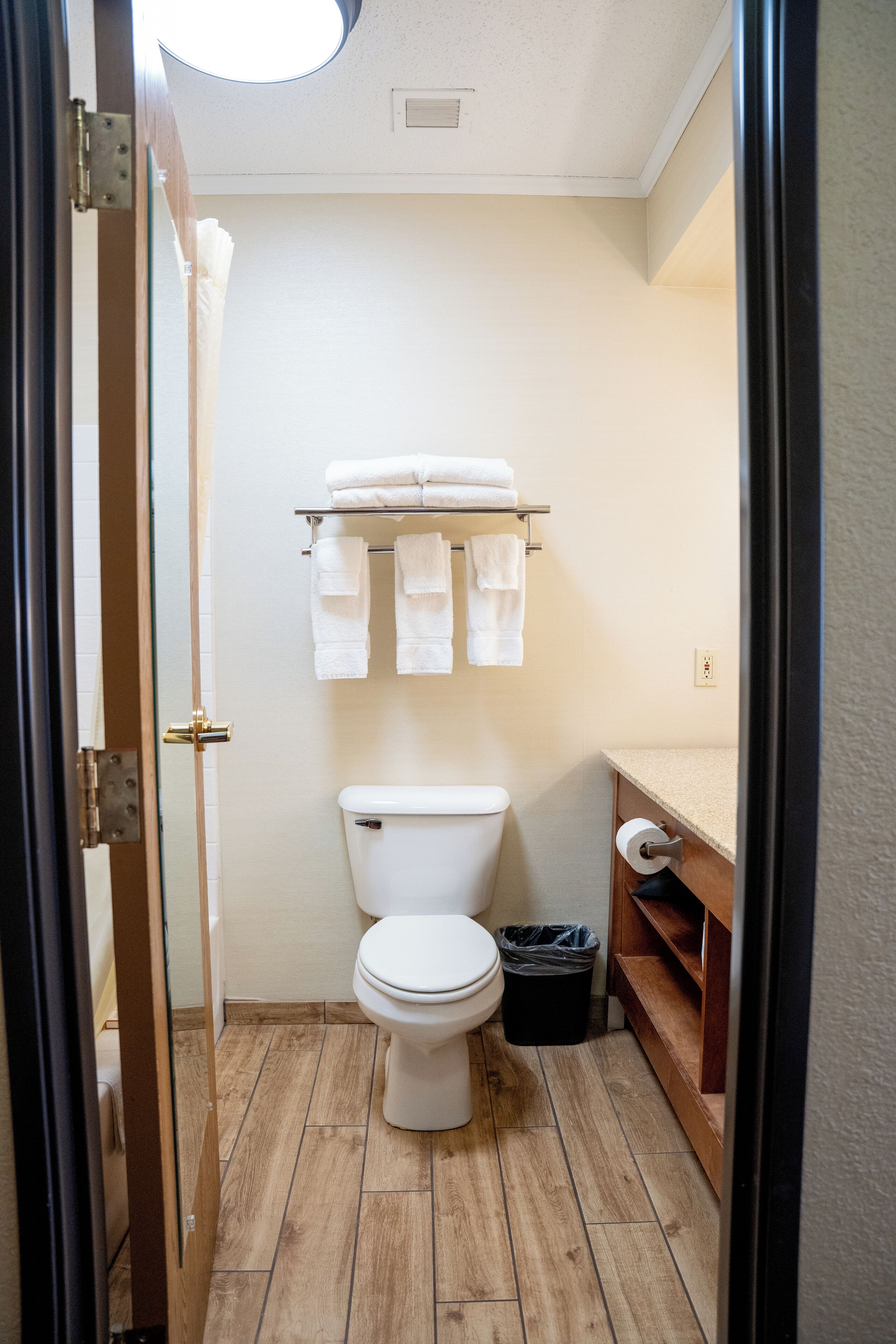 Een kleine badkamer met toilet, een plank met handdoeken, een houten vloer en een douchegordijn.
