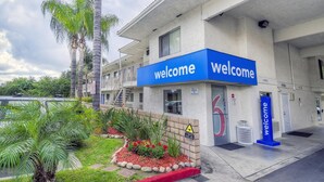 A building with a 'Welcome' sign, a gated entrance, and a landscaped area with palm trees and plants. - Motel 6 Chino, CA - Los Angeles Ontario (Chino)