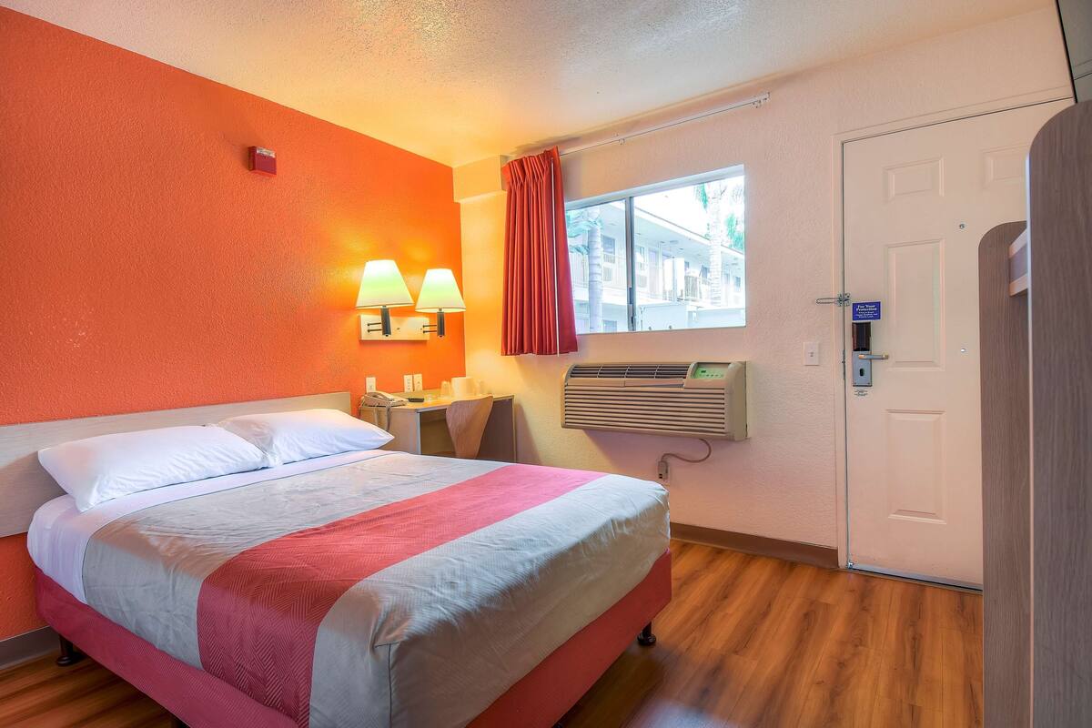 A hotel room with a bed, a desk, a chair, a window with curtains, and a door.