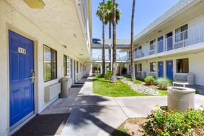 A hotel corridor with blue doors, a covered walkway, and a small garden area with palm trees and flowers. - Motel 6 Mesa, AZ - South (Mesa)