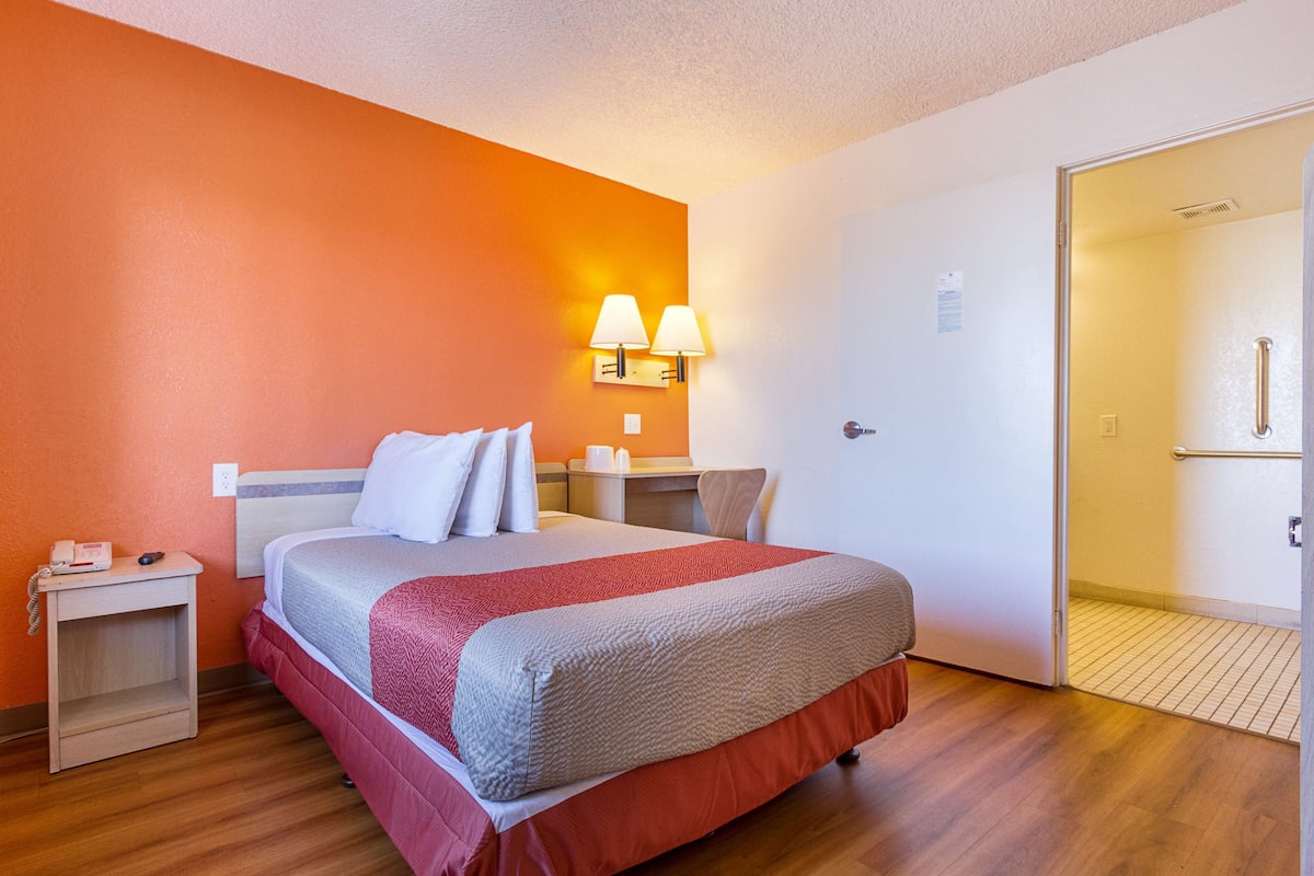 Una camera d'albergo con un letto, un comodino, una lampada a parete e una porta che dà su un bagno.