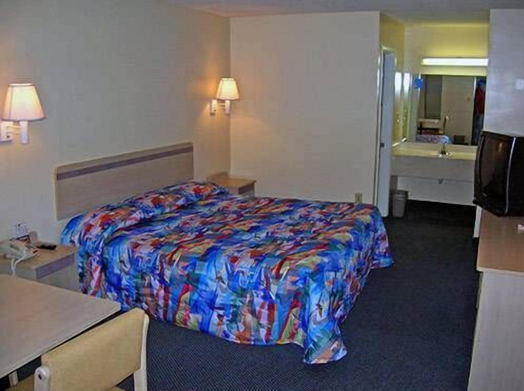 A hotel room with a bed, a desk, a chair, a lamp, and a mirror.