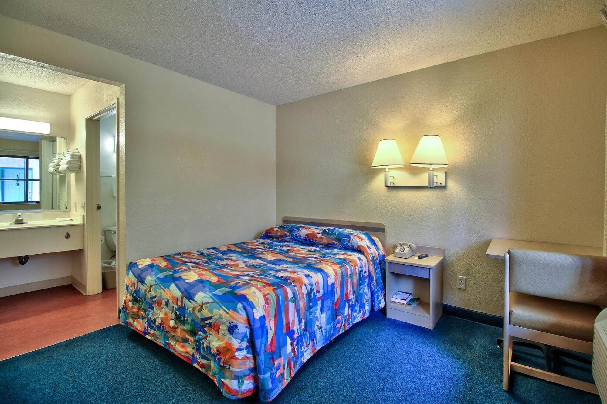 A hotel room with a bed, a nightstand, a desk, a chair, a bathroom with a mirror and sink, and two wall-mounted lamps.