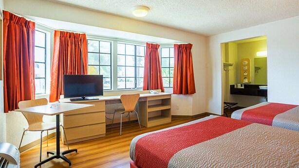 A hotel room with a desk, chair, TV, two beds, and a bathroom with a mirror. - Motel 6 Yuma, AZ - East (Yuma)