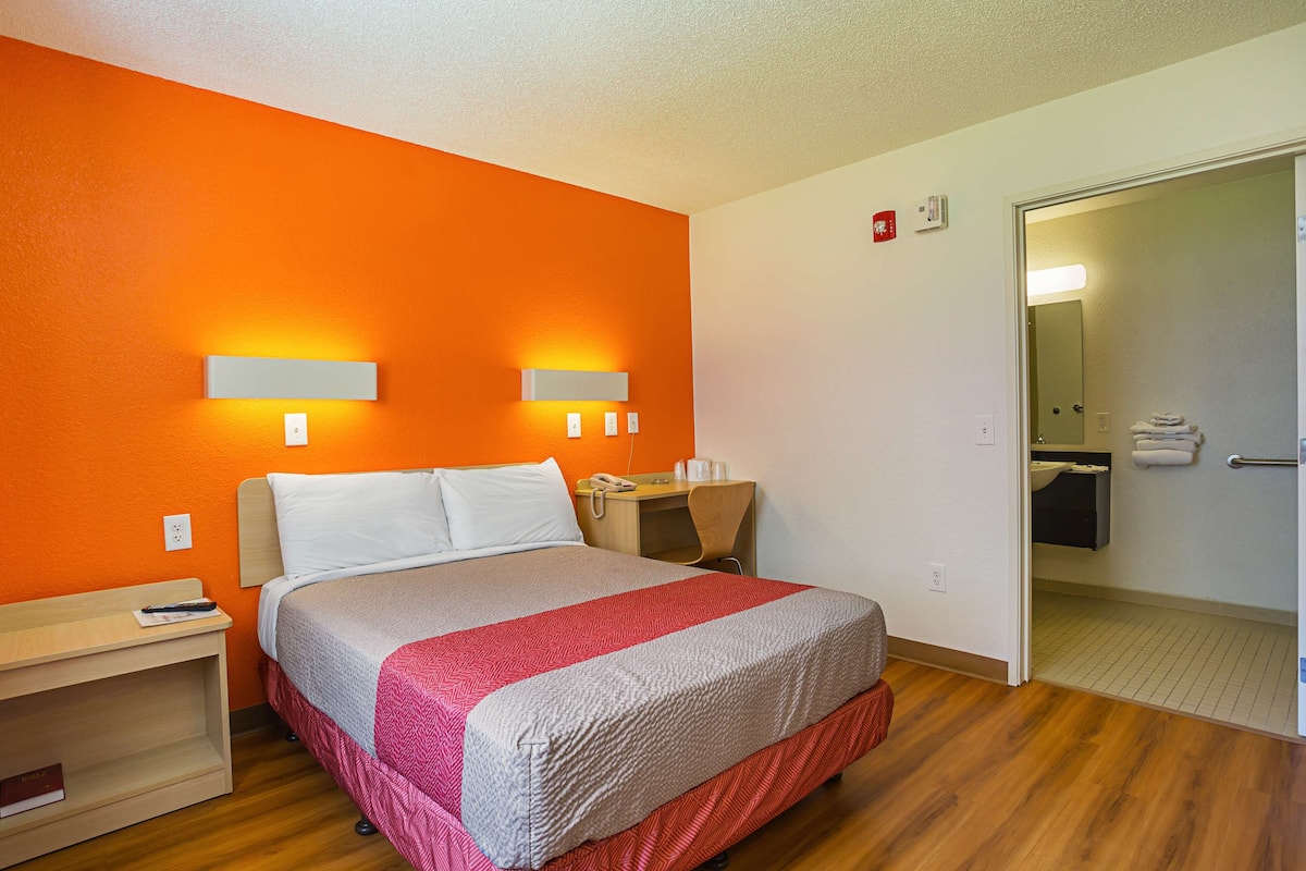 A hotel room with a bed, bedside table, and a desk with a chair. There is a bathroom visible through an open door.