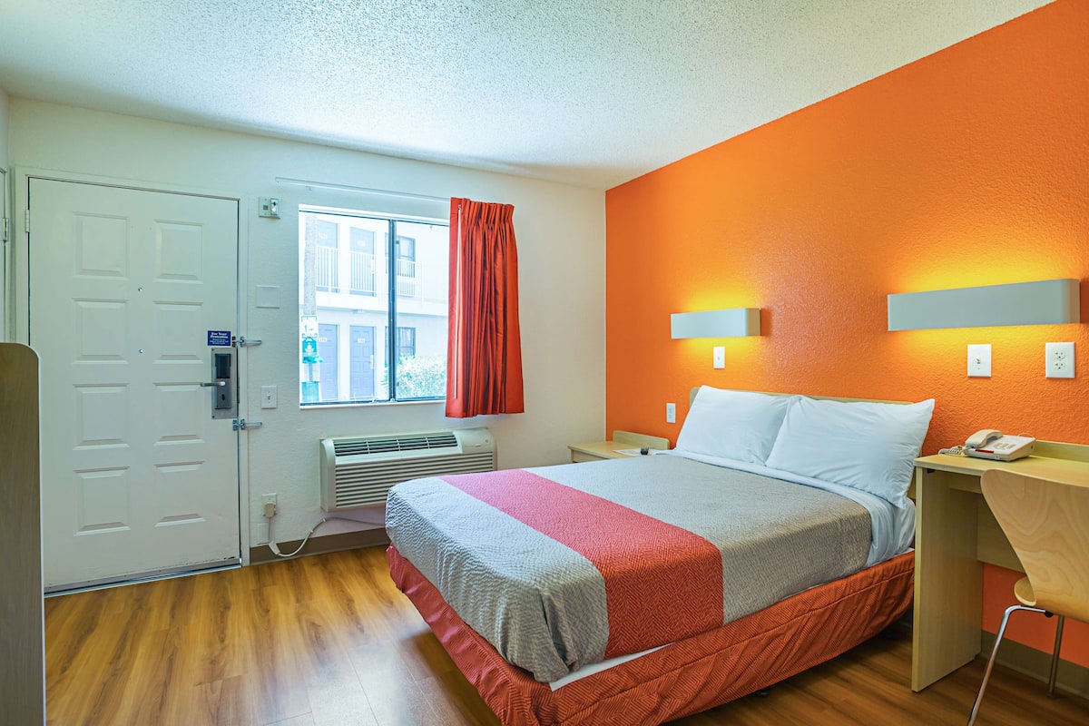 A hotel room with a bed, a desk, a chair, a window with curtains, and a door.